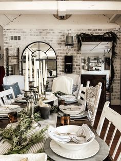 a dining room table set for christmas with white dishes and silverware on it, surrounded by greenery