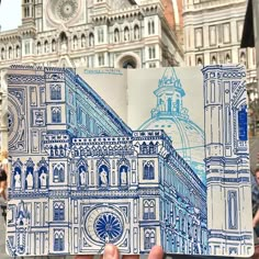 a hand holding up a blue and white drawing in front of a building with a clock tower