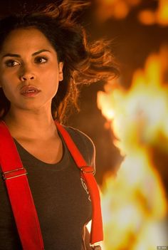 a woman in black shirt and red suspenders standing next to fire with her hair blowing in the wind