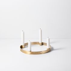 three white candles are placed in a brass ring on a white surface with one candle lit