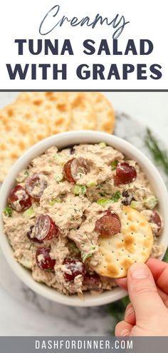 a hand holding a cracker over a bowl of tuna salad with grapes and cranberries