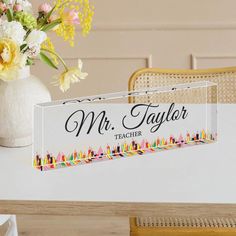 a teacher's name sign sitting on top of a table next to a vase with flowers