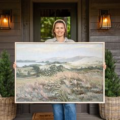 a woman holding up a large painting in front of a house with potted plants