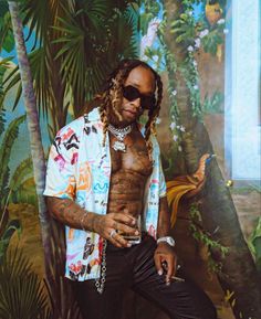 a man with dreadlocks sitting in front of a painting wearing a shirt and sunglasses