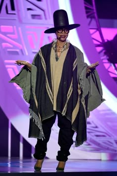 a man in a black hat is walking down the runway with his hands out and one hand on his hip