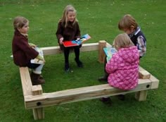 four children sitting on a wooden bench in the grass, reading books to each other