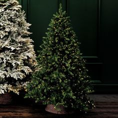 two artificial christmas trees sitting next to each other on top of a wooden floor in front of a green wall