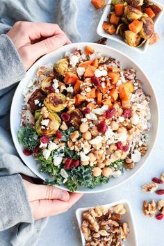 two hands holding a white bowl filled with vegetables and nuts next to another bowl full of food