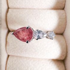 an engagement ring with two pear shaped diamonds and a pink diamond in the center on a white cushion