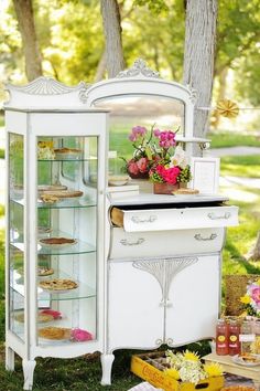 an old fashioned white cabinet with flowers and cakes in it's glass doors is sitting on the grass