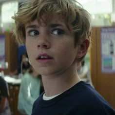 a young boy looks at the camera while standing in an office with other people behind him