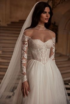 a woman wearing a wedding dress with long sleeves and a veil on her head standing in front of stairs