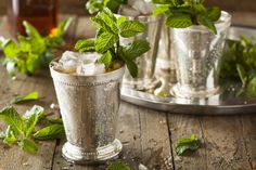 a glass filled with ice and mint sitting on top of a wooden table next to two silver cups