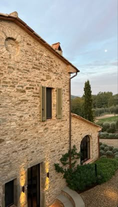 an old stone building with green shutters on the front and side windows, surrounded by greenery