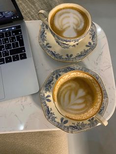 two cups of coffee sit next to each other on a table with an open laptop