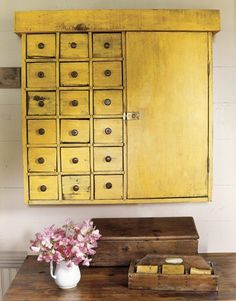 an old yellow cabinet with drawers and flowers in it next to a vase on a wooden table