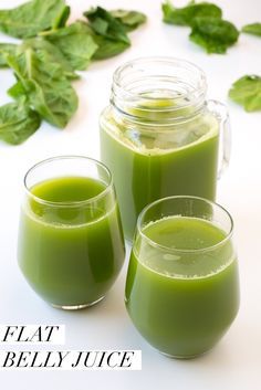 two glasses filled with green liquid next to some leaves on a white surface and another glass full of juice