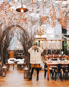 a woman standing in front of a table with chairs and trees hanging from the ceiling