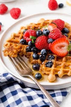 a waffle topped with berries and blueberries on a white plate next to a fork