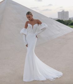 a woman in a white wedding dress standing on the sand with her arms behind her back