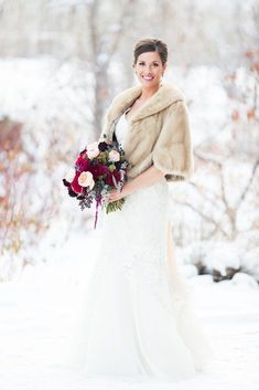 a woman in a wedding dress holding a bouquet and fur stole over her shoulders while standing in the snow