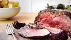 a large piece of meat sitting on top of a wooden cutting board next to a bowl of potatoes