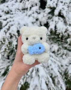 a hand holding a small teddy bear in front of snow covered trees and evergreens