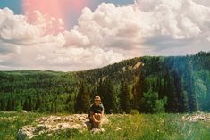 a man sitting on top of a lush green hillside under a colorful sky with clouds