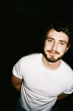 a man with a goatee and beard standing in the dark wearing a white t - shirt