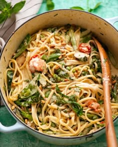 a pot filled with pasta and vegetables on top of a green table cloth next to a wooden spoon