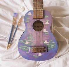 a purple ukulele sitting on top of a bed next to a paintbrush