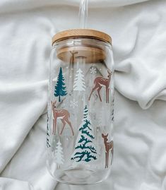 a glass jar with deer and pine trees painted on the side, sitting on a white blanket