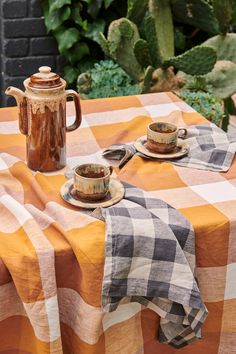 an orange and white checkered table cloth with two cups on it, next to a potted plant