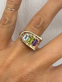 This one caught my eye immediately and I just knew I had to try it on.  It feels just as awesome on the finger as it looks.  It sits very low to the finger with excellent finger coverage. It golds 3 genuine emerald cut gemstones: Amethyst, Peridot, and Aquamarine.  Each stone measures 6mm x 4mm.   The ring has a nugget-style detail on one shoulder of the ring while the other shoulder is high-polish. The ring weighs 7.8g and measures 12mm at its widest point. The ring tapers to 3mm at the back of the shank. Ring is not stamped but guaranteed to be 14K yellow gold. The ring is currently a size 6.25 and sizable.  We are happy to provide sizing for an additional charge. Please let me know if you need a quote. Modernist Ring, Emerald Cut, Rings Statement, Band Ring, Aquamarine, Band Rings, Statement Rings, Emerald, Jewelry Rings