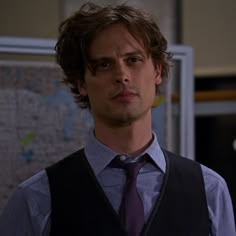 a man in a vest and tie looking at the camera with a map on the wall behind him