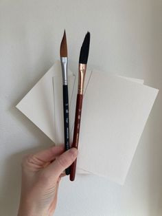 a hand holding three brushes next to two blank cards and envelopes on a white surface