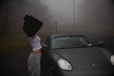 a woman standing next to a sports car in the fog with her hand on the hood