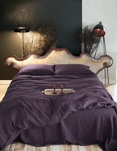 a bed with purple sheets and pillows in a room next to a lamp on the wall