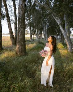 a woman in a white dress is standing by some trees and holding a pink bouquet