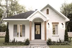 a white house with black door and windows on the front porch is surrounded by greenery