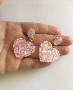 a hand holding two pink heart shaped earrings in it's palm, with glitter on them