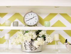 a shelf with flowers, candles and a clock on it in front of a chevron wall