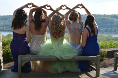 four women in dresses sitting on a bench making heart shapes with their hands while looking at the water