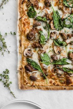 a square pizza with mushrooms, spinach and cheese on a white plate next to some herbs