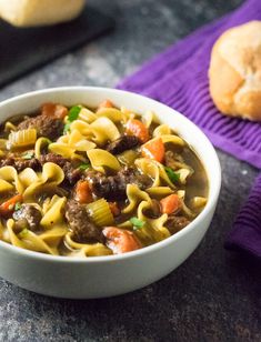 a white bowl filled with pasta and meat soup on top of a purple towel next to rolls