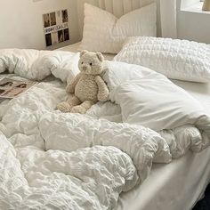 a teddy bear sitting on an unmade bed with white comforters and pillows in a bedroom