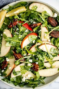 a salad with apples, spinach and pecans in a white bowl on a marble table