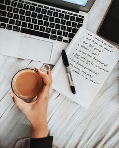 a person holding a cup of coffee in front of a laptop and notepad on a bed