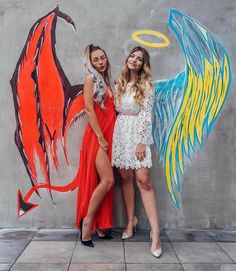 two beautiful women standing next to each other in front of a wall painted with wings