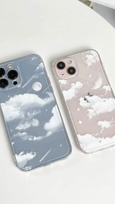 two iphone cases sitting next to each other on top of a white table with clouds in the sky